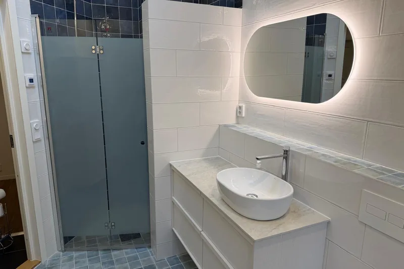 Badrum renoverat av BHB Bygg AB med vitt kakel, blå mosaik, belyst oval spegel och duschvägg.